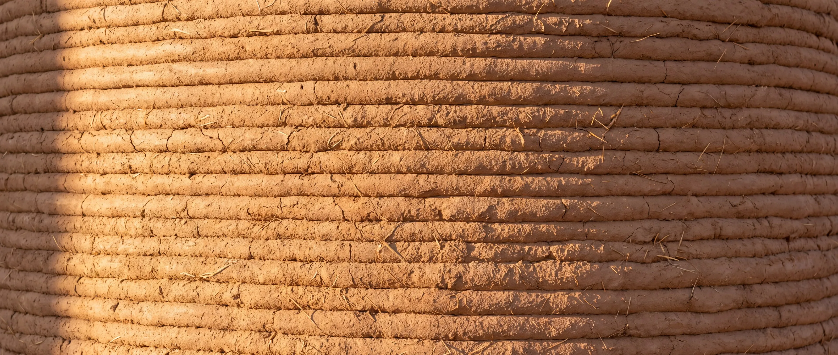 Close-up of a curved wall of 3D printed earth: stacked beads with straw fibres and fine cracks, lit from the side.