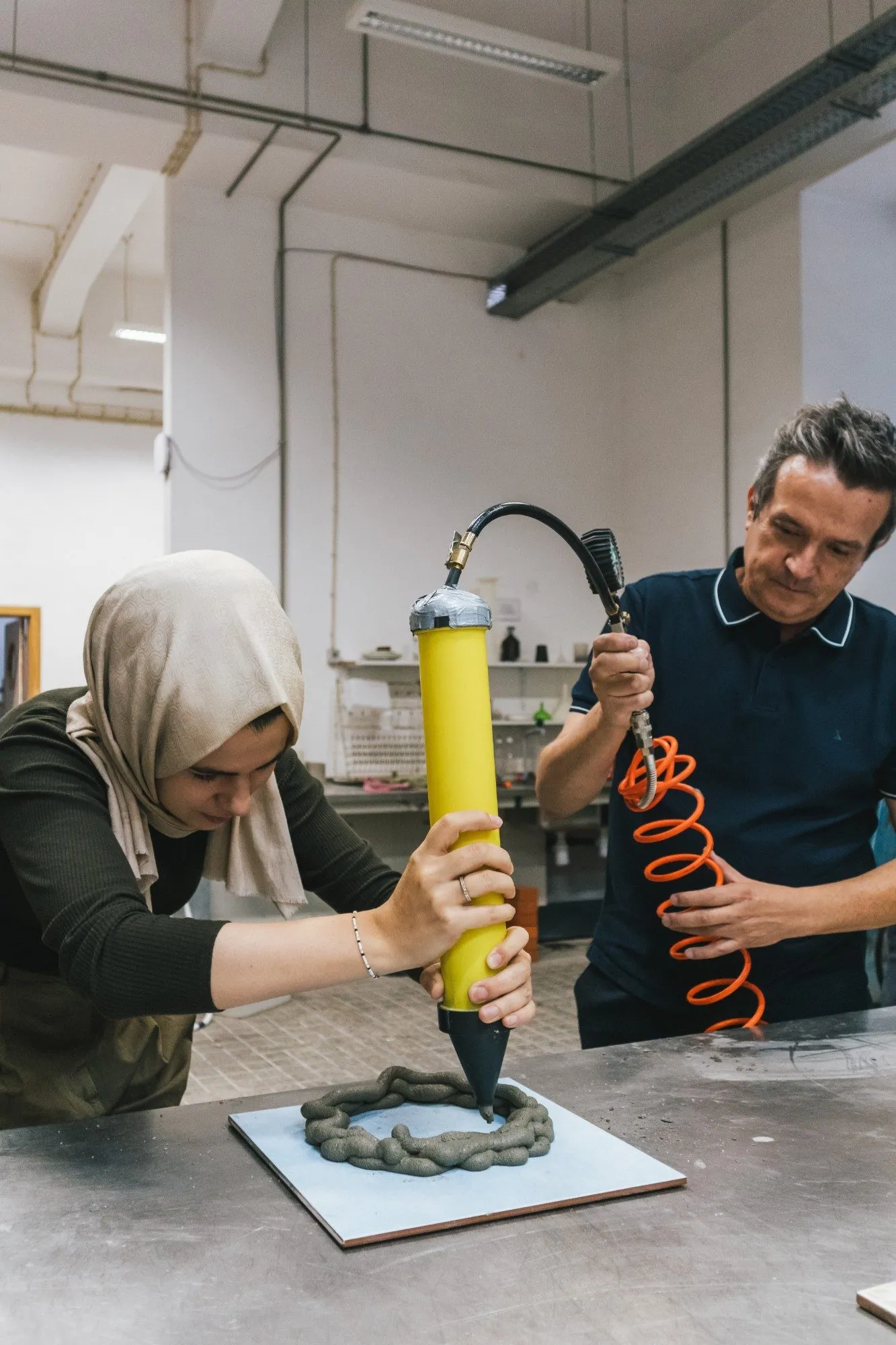 A participant extrudes a coil of material from a yellow cartridge onto a board, building it up layer by layer, while another participant holds the air hose.