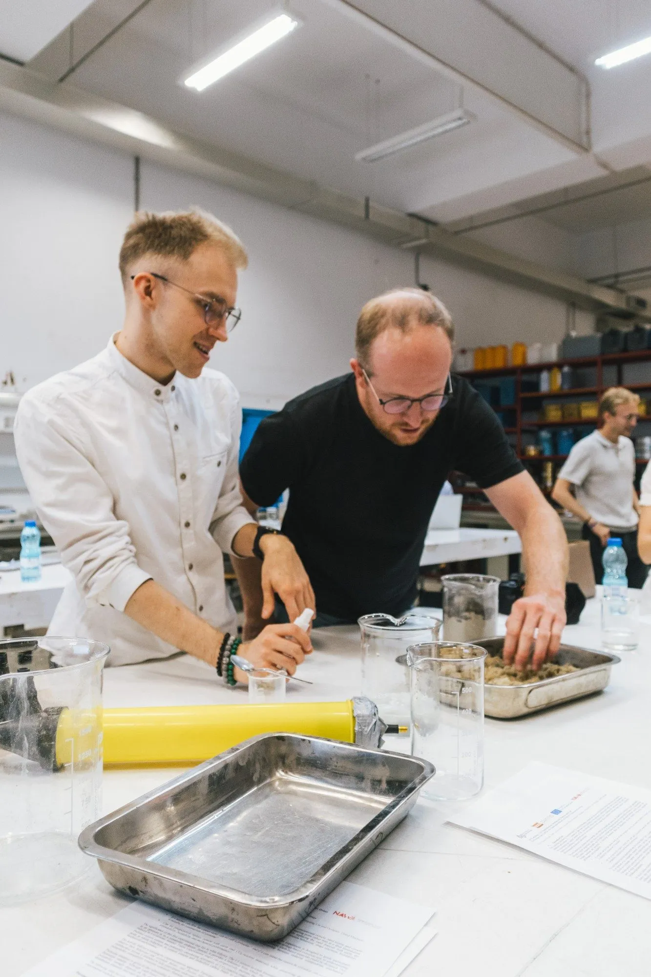 Two participants prepare a material mix in a metal tray at a laboratory bench, next to a yellow extrusion cartridge.