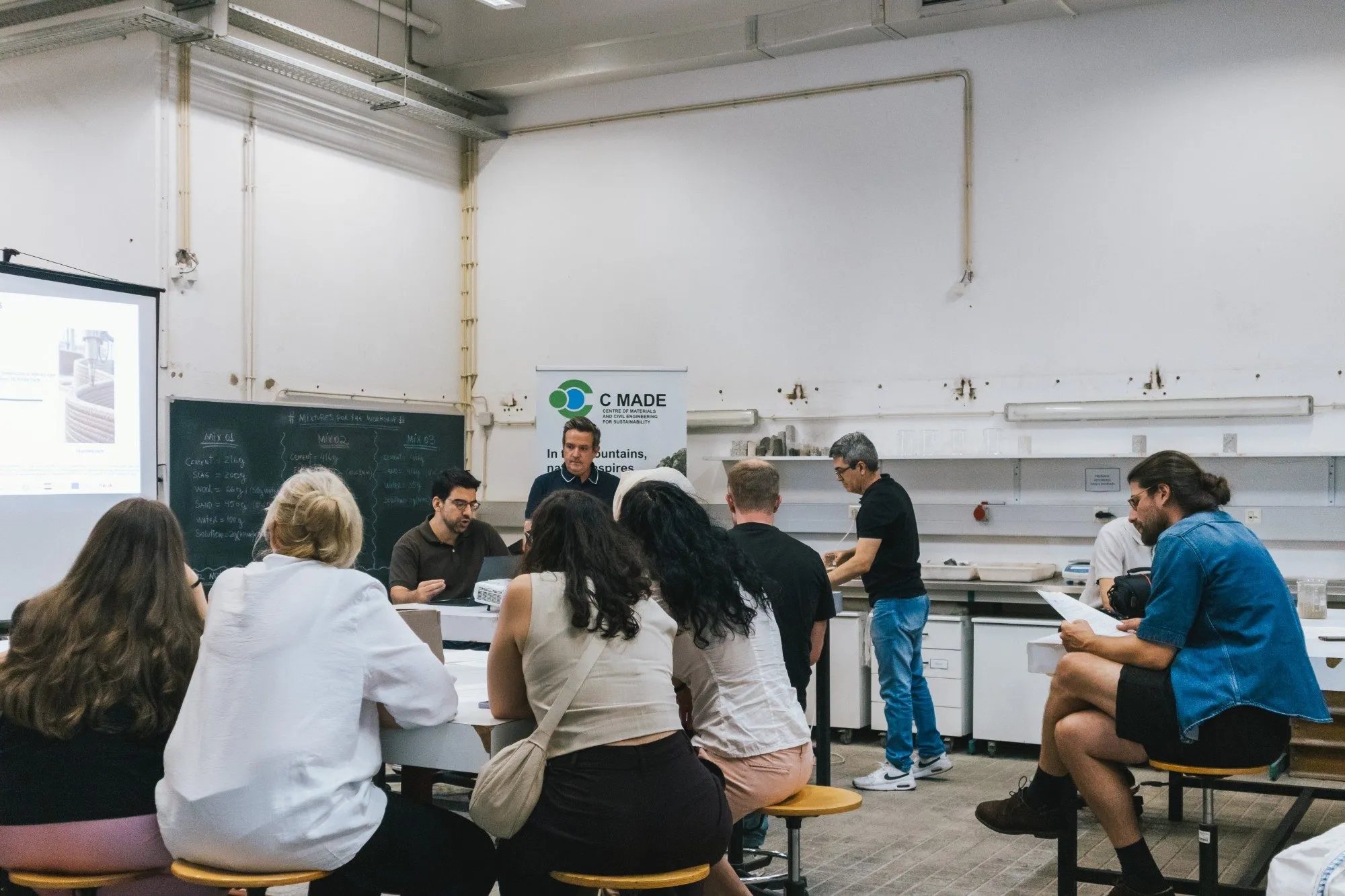 Participants on laboratory stools listen to a speaker at the front of a university laboratory, next to a C MADE banner.
