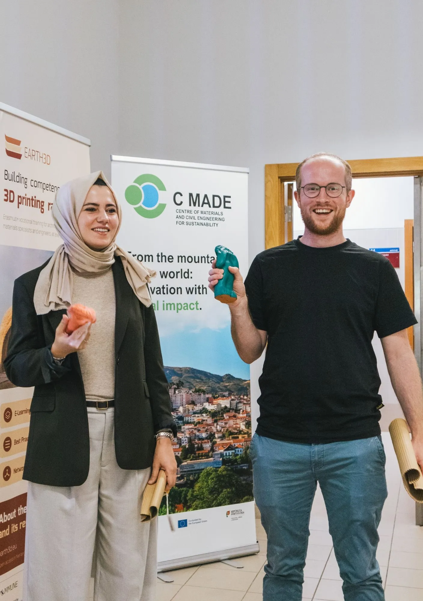 Two participants smile in front of the EARTH3D and C MADE banners, each holding a small printed object.
