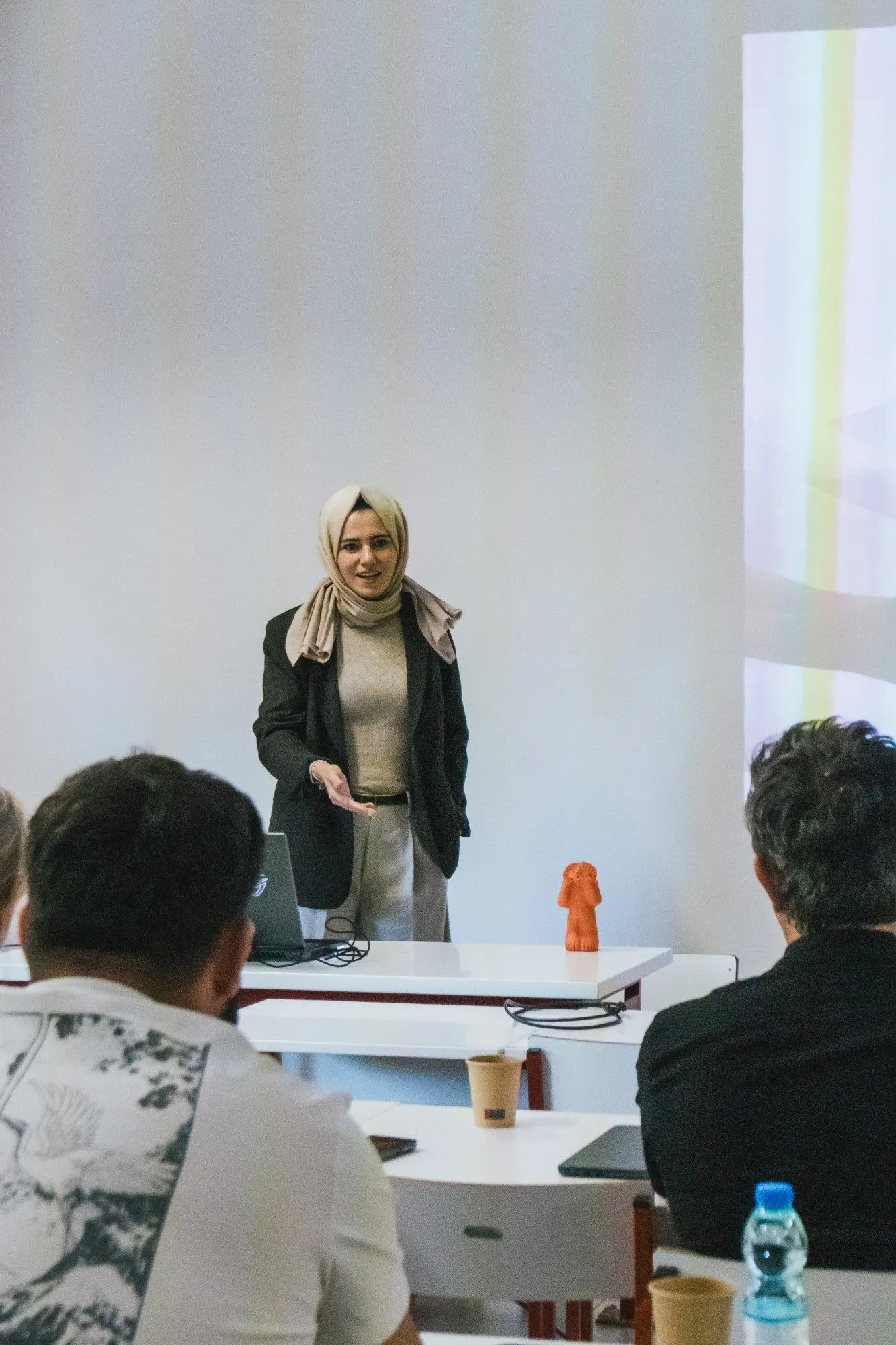 A speaker stands in front of participants seated at tables in a classroom.