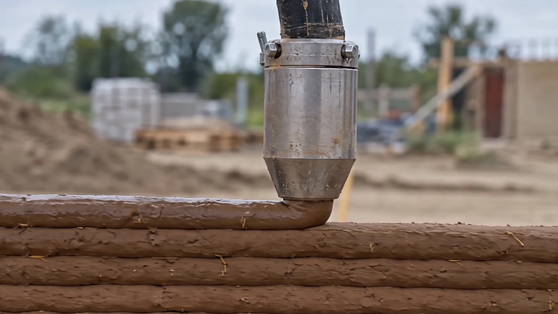 A steel nozzle lays a bead of raw earth on top of the earlier layers of a printed wall, on an outdoor building site.