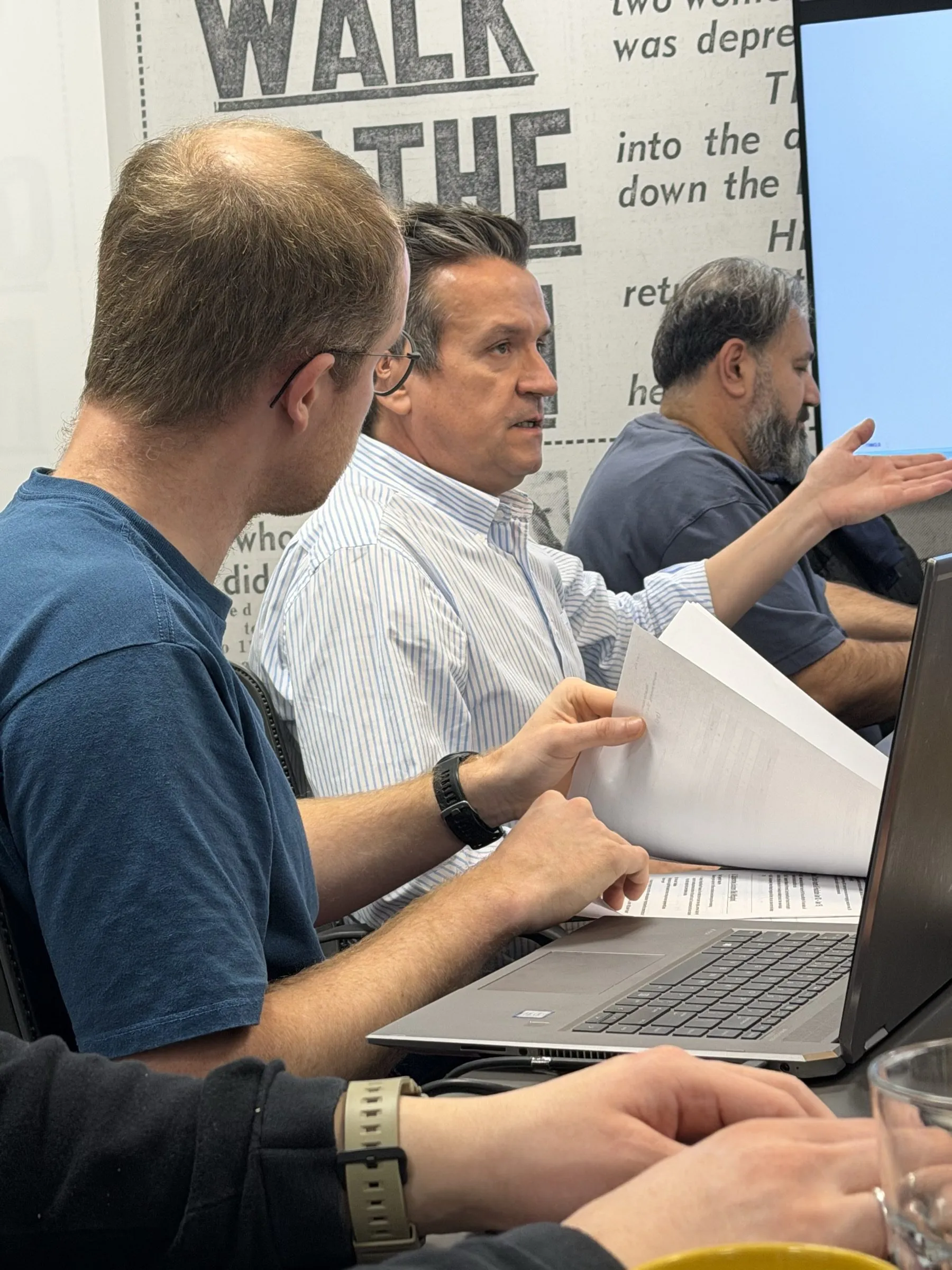 Partners in discussion at the meeting table, one of them holding printed documents.